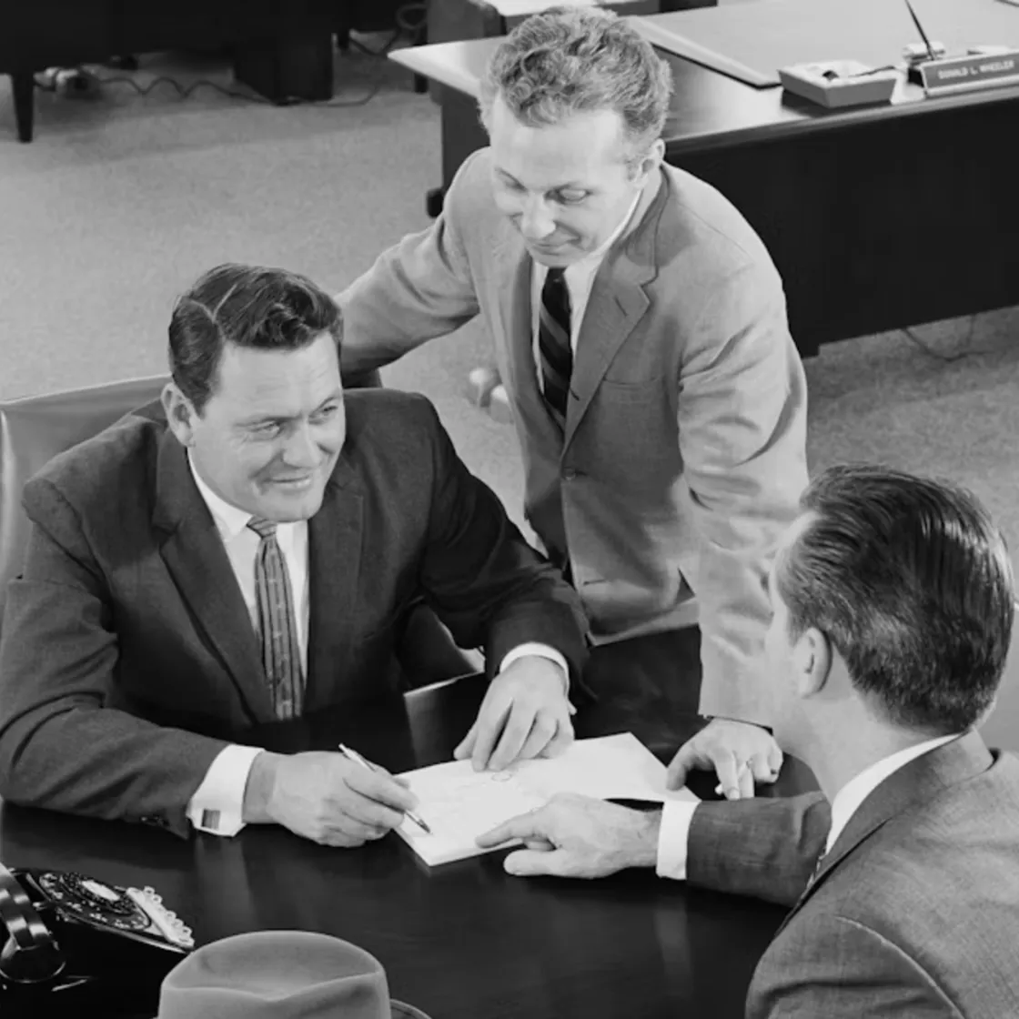 Three businessmen in suits finalizing a deal at an office desk, circa 1960.